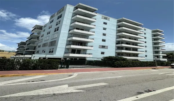 Multi-story residential building featuring a light blue stucco exterior, stacked private balconies with black metal railings, and a gated ground-level parking entrance