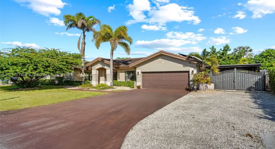Single-story residence featuring a stucco exterior, an attached two-car garage, an asphalt driveway, a gravel parking area, and decorative metal gates