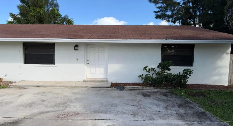 Exterior featuring a white stucco facade, brown shingle roof, concrete driveway, and exterior wall-mounted lighting
