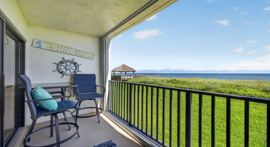 Covered balcony featuring concrete flooring and black metal railing, overlooking a landscaped green space and coastal water