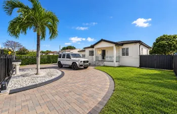 Single-story exterior featuring a paver driveway, covered front entryway, white stucco facade, perimeter fencing, and established palm tree landscaping