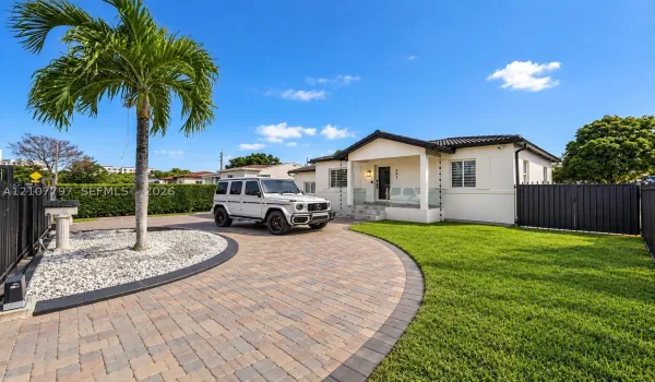 Single-story exterior featuring a paver driveway, covered front entryway, white stucco facade, perimeter fencing, and established palm tree landscaping