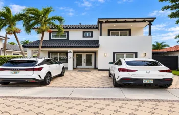 Two-story exterior featuring a high-contrast stucco facade, dark roof tiles, a covered entry with French doors, a second-level balcony, and an expansive paved driveway