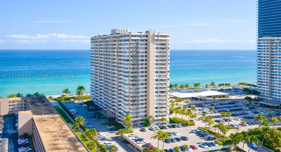 High-rise residential building featuring stacked private balconies, an expansive paved parking area, and mature palm tree landscaping