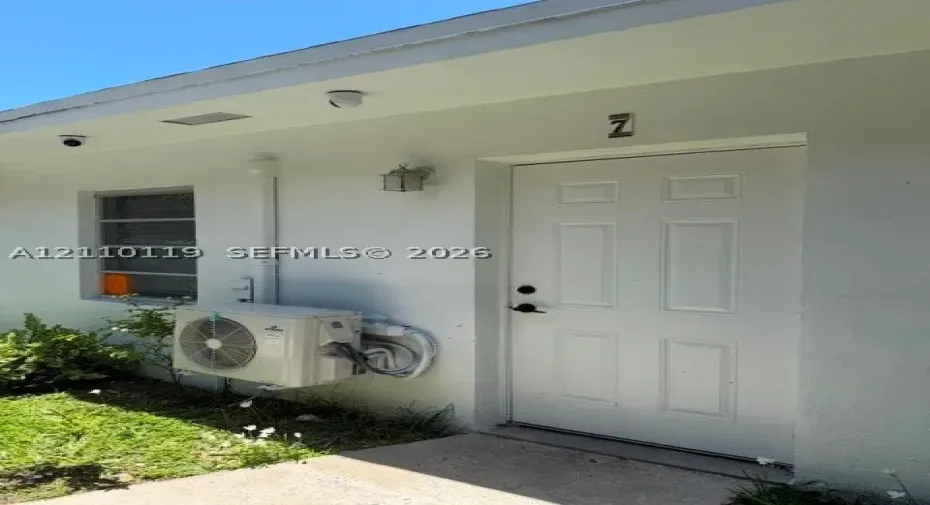 Exterior entrance featuring a white panel door, metal-framed window, wall-mounted carriage-style light fixture, and an externally mounted ductless HVAC unit