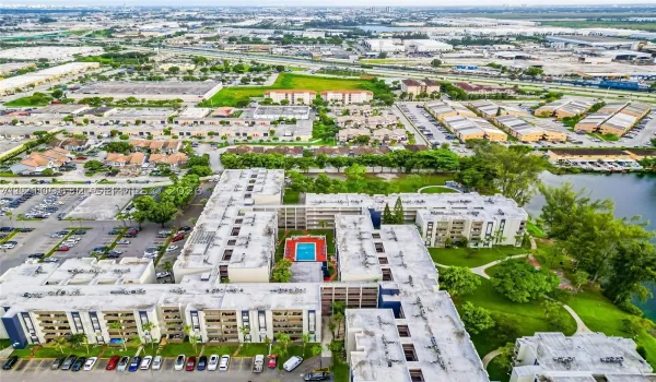 Aerial perspective of a multi-story residential complex featuring a central swimming pool, paved surface parking, manicured green space, and proximity to a canal