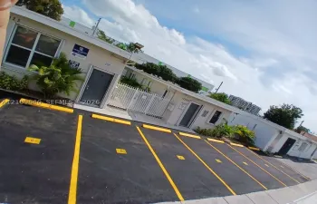 Paved parking area with marked stalls and wheel stops, exterior building facade with stucco finish, white metal security fencing, and pedestrian entry doors