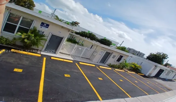 Paved parking area with marked stalls and wheel stops, exterior building facade with stucco finish, white metal security fencing, and pedestrian entry doors