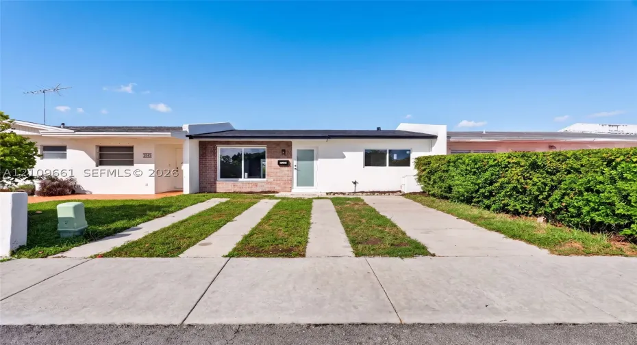 Single-story exterior featuring white stucco siding, brick masonry detailing, horizontal window slats, and a concrete ribbon driveway