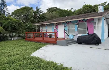 Single-story residence featuring an exterior mural, raised wood-finish deck with railing, shingle roof, concrete driveway, and mature landscaping