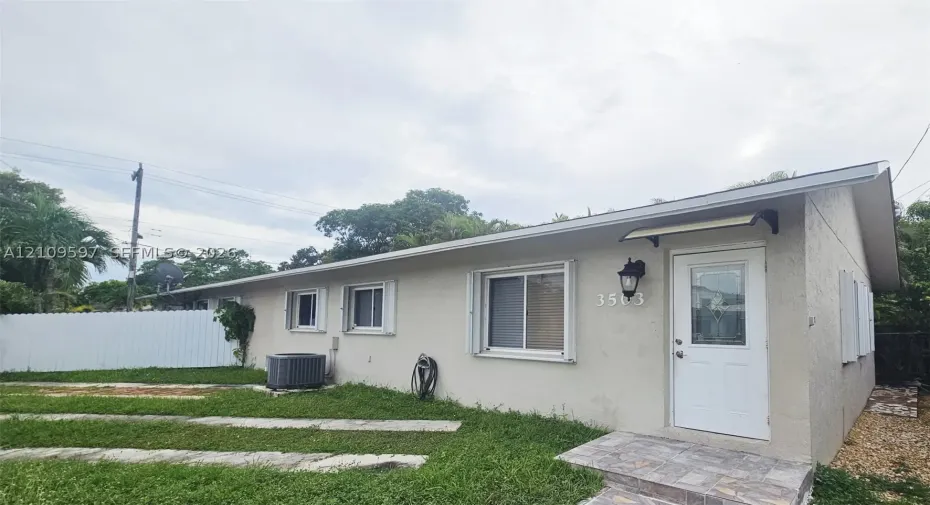 Single-story exterior featuring light-colored stucco siding, a shingled roof, sliding windows with white frames, a white entry door with a decorative glass inset, and stone-finish porch tiles