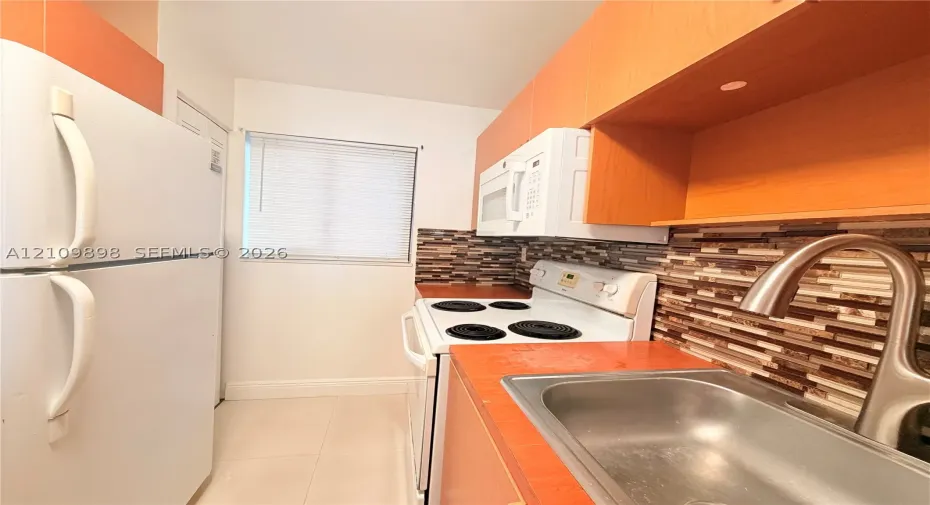 Kitchen featuring wood-tone cabinetry, a decorative tile backsplash, white appliances, and a stainless steel sink with a high-arch faucet