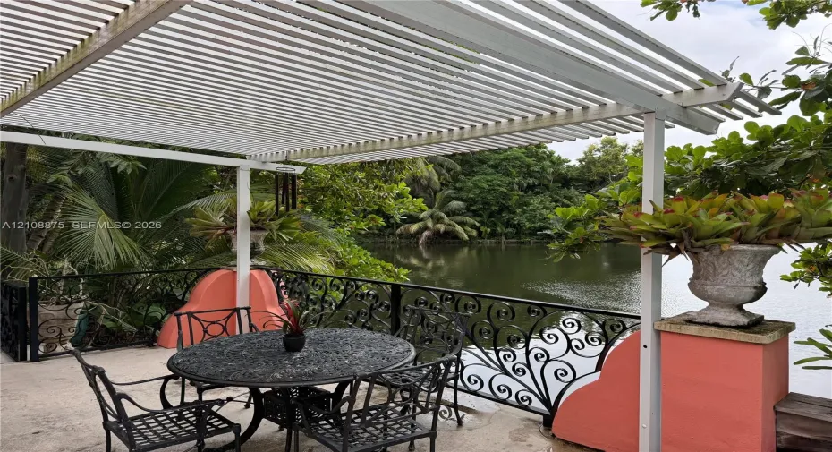 Outdoor patio featuring a white louvered pergola, black wrought-iron railing, concrete flooring, and decorative masonry pillars