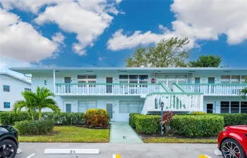 Two-story building exterior featuring a second-level balcony with white balustrades, horizontal window configurations, exterior stair access, and manicured shrubbery