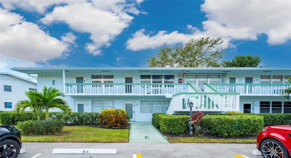 Two-story building exterior featuring a second-level balcony with white balustrades, horizontal window configurations, exterior stair access, and manicured shrubbery