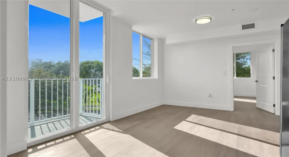 Floor-to-ceiling glass sliding doors, private balcony with white railing, wood-finish flooring, and recessed ceiling lighting