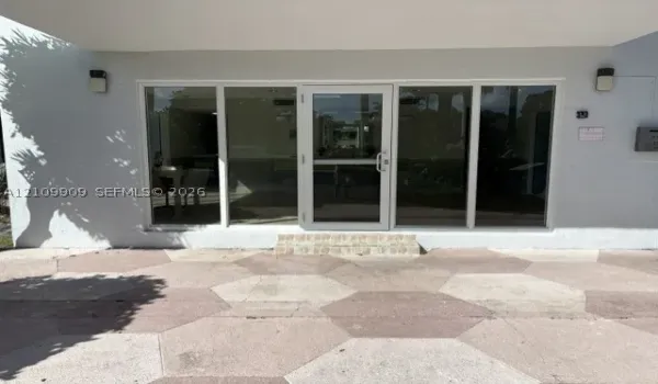 Building entrance featuring a multi-pane glass door, full-height glass side panels, textured exterior walls, and a hexagonal paver entryway