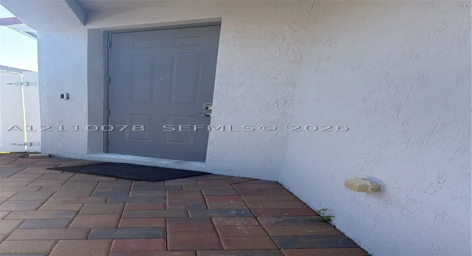 Exterior entryway featuring a textured stucco finish, multi-panel gray door, and brick-style paver flooring