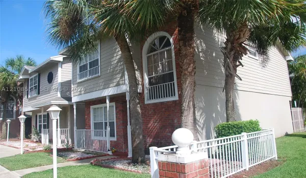 Multi-story exterior featuring a red brick and horizontal siding facade, arched second-story windows, metal balcony railings, and mature palm trees