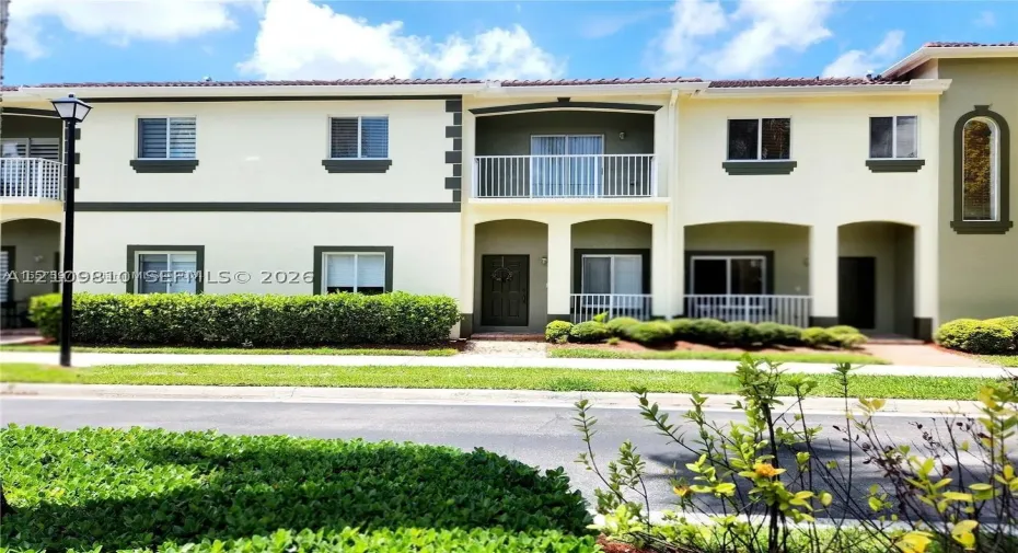 Two-story exterior featuring light-toned stucco walls with contrasting trim, a tiled roof, a private upper-level balcony with white railings, and covered lower-level entryways
