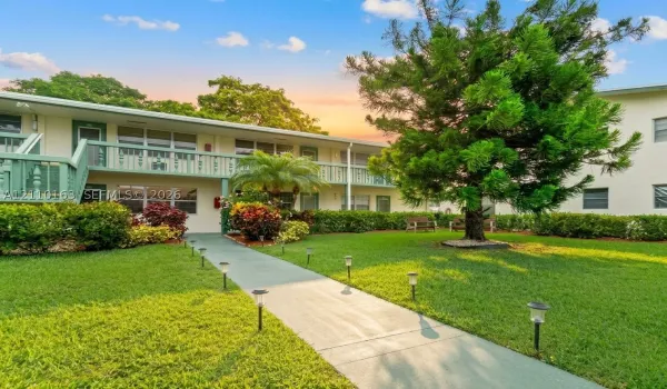 Two-story exterior featuring painted stucco facade, second-level balcony with railings, concrete pedestrian walkway, manicured lawn, and mature landscaping