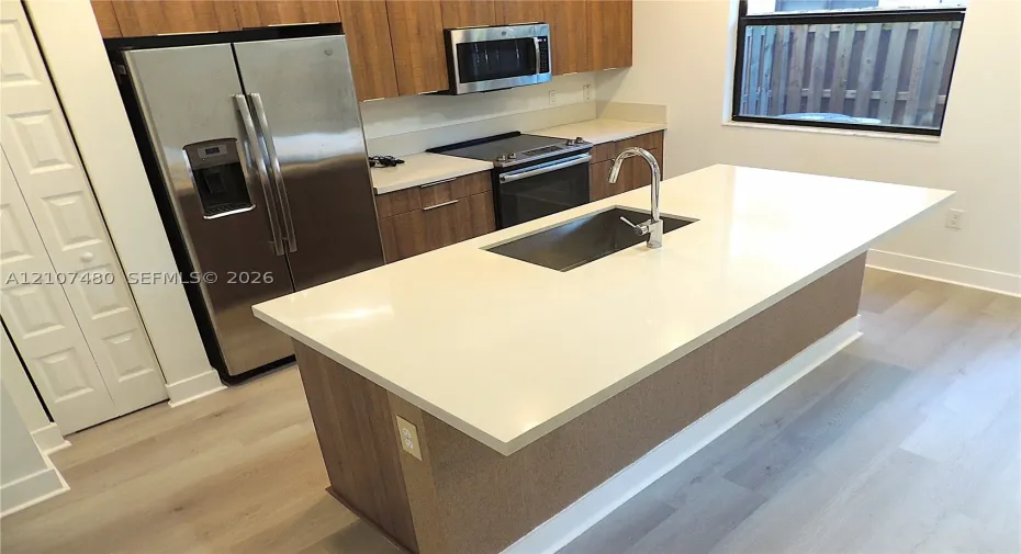 Kitchen featuring a central island with a solid-surface countertop and undermount sink, stainless steel appliances, flat-panel wood-finish cabinetry, and wood-finish flooring