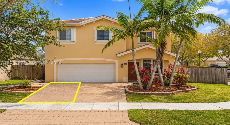 Two-story stucco exterior featuring a paved stone driveway, an integrated two-car garage, and landscaped garden beds with mature palms
