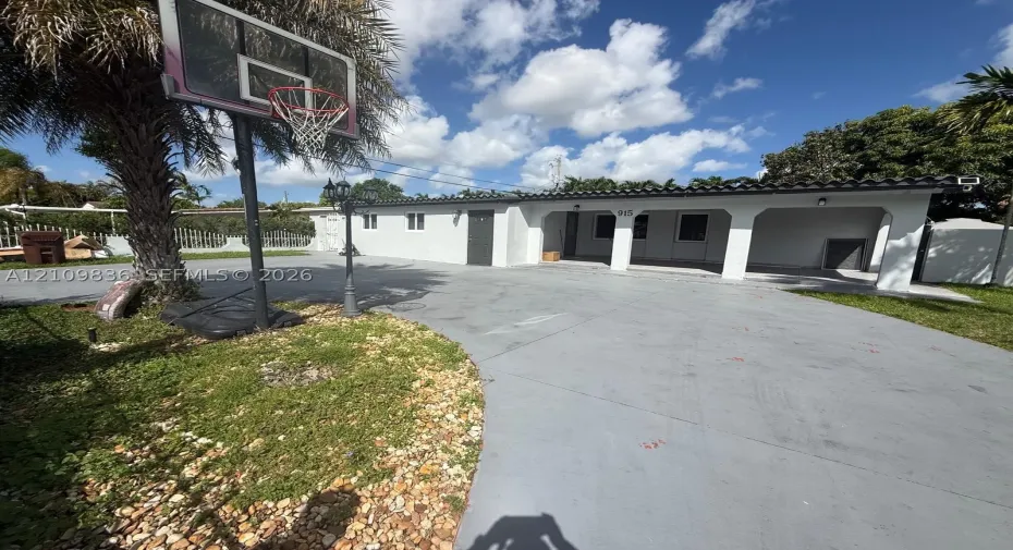 Exterior facade featuring a covered carport, smooth stucco finish, barrel-tile roofline, and an expansive paved driveway