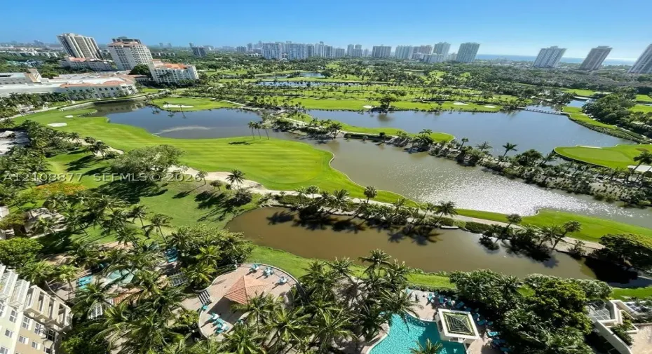 Aerial perspective featuring a large swimming pool, paved pool deck, and a pavilion structure, overlooking expansive green space, multiple water features, and a skyline horizon