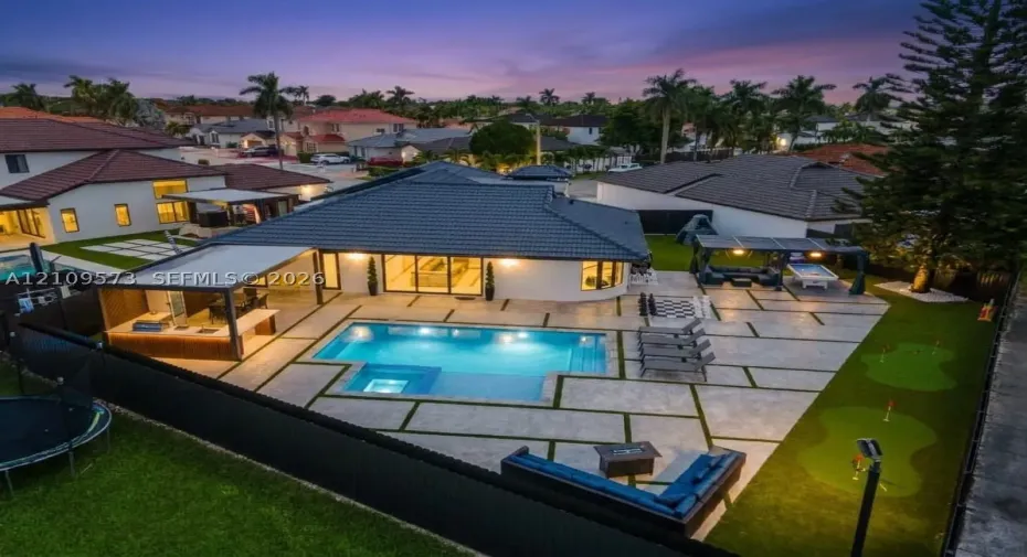 Backyard area featuring an in-ground pool with integrated spa, concrete paver decking with grass inserts, covered outdoor kitchen, and synthetic turf putting green