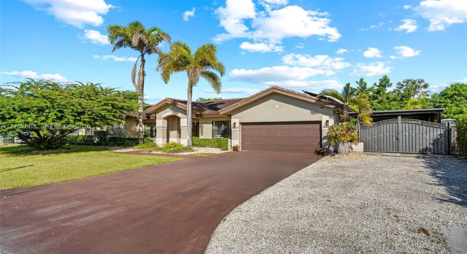 Single-story residence featuring a stucco exterior, an attached two-car garage, an asphalt driveway, a gravel parking area, and decorative metal gates