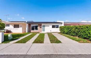 Single-story exterior featuring white stucco siding, brick masonry detailing, horizontal window slats, and a concrete ribbon driveway