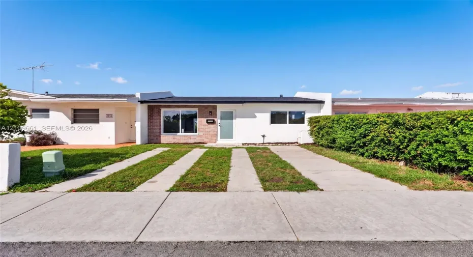 Single-story exterior featuring white stucco siding, brick masonry detailing, horizontal window slats, and a concrete ribbon driveway