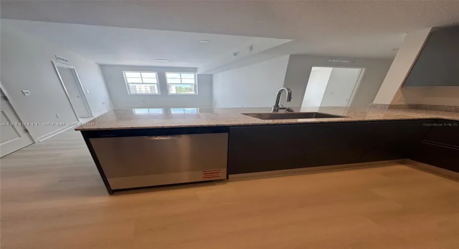 Kitchen featuring a stone-composite countertop, stainless steel dishwasher, undermount stainless steel sink with a high-arch faucet, flat-panel dark cabinetry, and light-toned wood-finish flooring