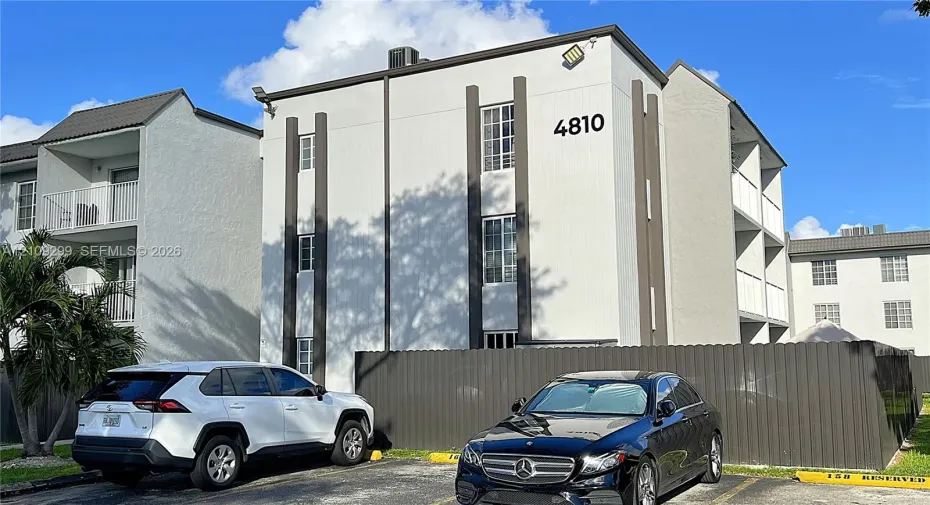 Three-story multi-family structure featuring a smooth stucco exterior, vertical architectural accents, grid-pattern windows, and a metal perimeter fence