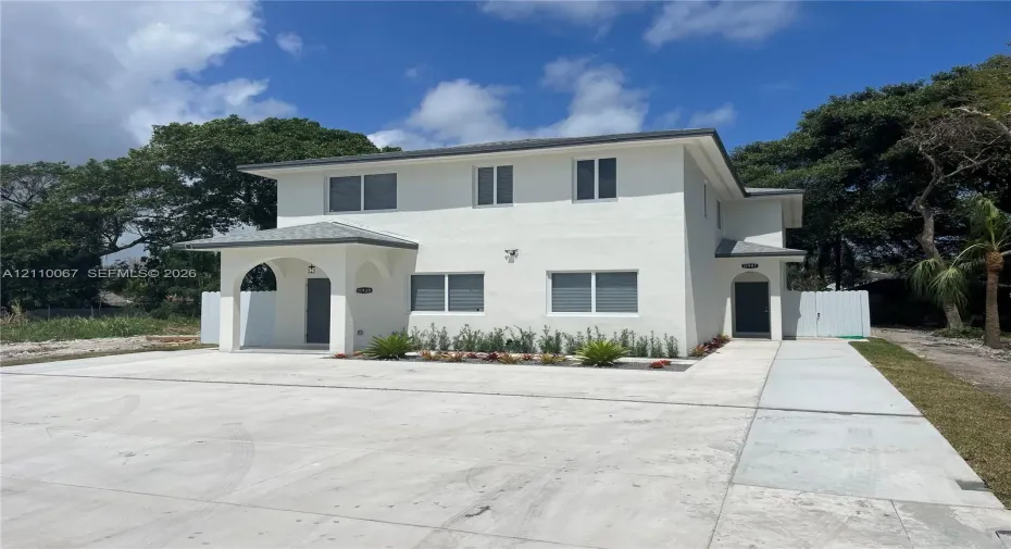 Two-story exterior featuring a white stucco facade, arched covered entryway, hip roofline, concrete driveway, and installed privacy fencing
