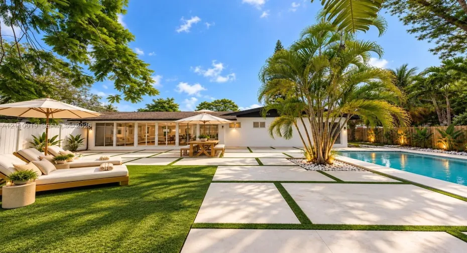 Rear exterior featuring a swimming pool, large-format paver patio with turf inlays, mature palm trees, and full-height glass sliding doors