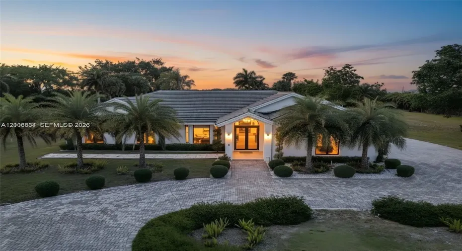 Single-story residence with a concrete tile roof, double-door entryway, and paver driveway, set on a landscaped lot with mature palm trees