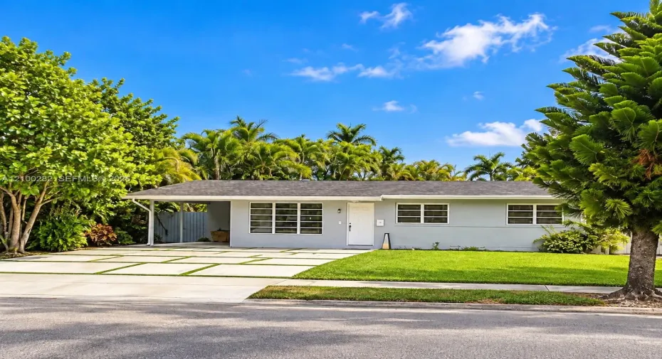 Single-story exterior featuring a light gray facade, covered carport, ribbon driveway with integrated greenery, multi-pane windows, and mature palm landscaping