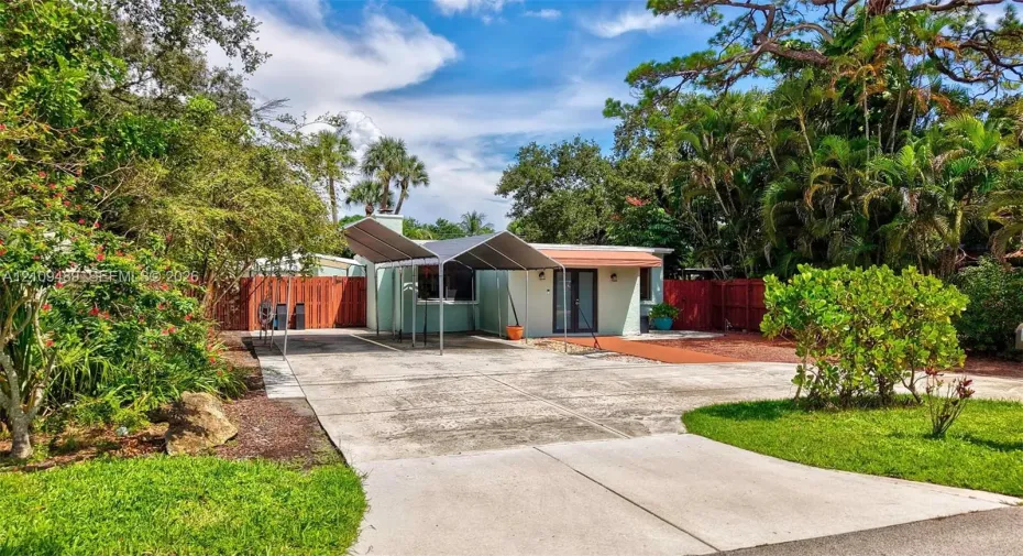 Concrete driveway with a metal carport, horizontal wood-style fencing, and mature landscaping