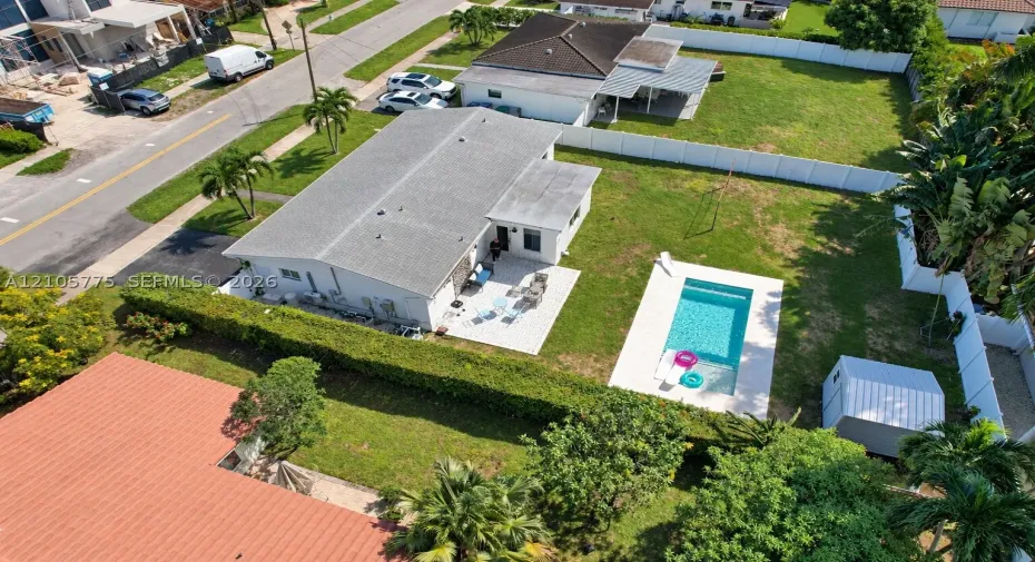 Aerial perspective of a property featuring an in-ground swimming pool, paved patio, expansive lawn, white vinyl perimeter fencing, and a detached storage structure
