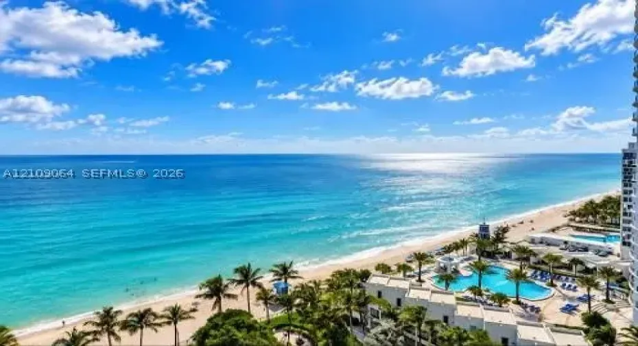 High-altitude perspective featuring an expansive ocean horizon, sandy coastline, and a multi-pool aquatic complex with surrounding palm trees