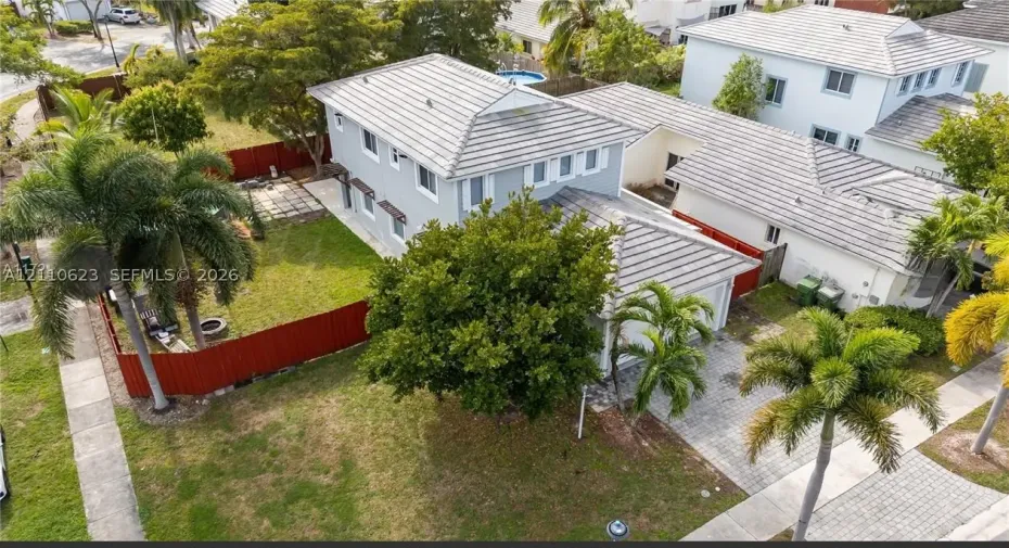 Two-story residence featuring a concrete tile roof, paver driveway, fenced yard, and established tropical landscaping