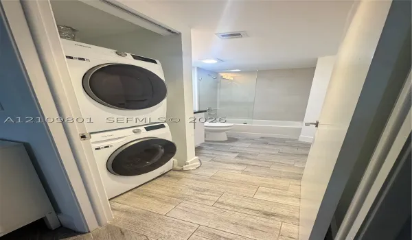 Stacked white appliance suite featuring a front-loading washer and dryer, complemented by wood-finish tile flooring, a bathtub with a glass panel, and neutral wall tile surrounds