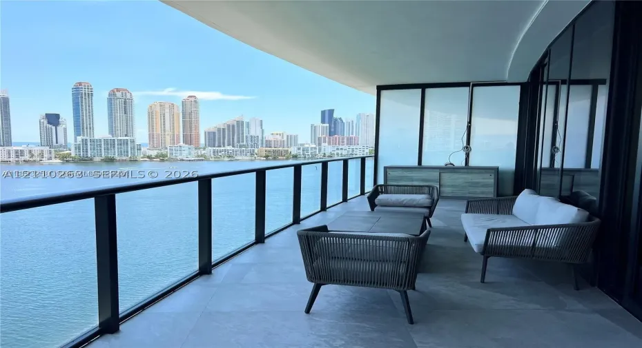 Expansive covered balcony featuring large-format stone flooring, black metal railing, and floor-to-ceiling glass wall panels