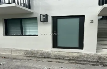 Exterior entrance featuring a white stucco facade, a black-framed door with a frosted glass panel, a sleek rectangular wall-mounted light fixture, and a black-framed window