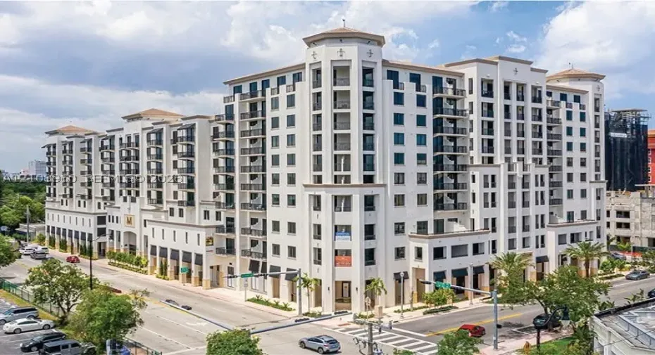 Mid-rise building featuring a light-colored stucco exterior, decorative corner turrets, tiered balconies with wrought-iron railings, and ground-level retail awnings
