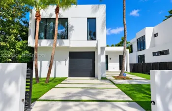 Modern two-story architectural exterior featuring smooth white stucco finish, large dark-framed windows, a sleek charcoal-colored garage door, and a geometric concrete paver driveway