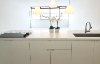 Kitchen island featuring a solid-surface countertop, integrated flat-surface cooktop, undermount stainless steel sink with a high-arch gooseneck faucet, minimalist flat-panel cabinetry, and three pendant lights