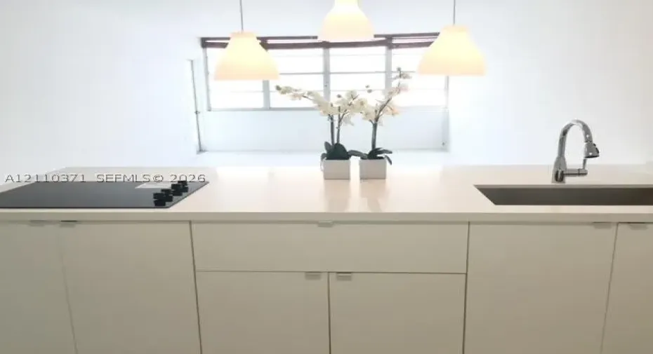 Kitchen island featuring a solid-surface countertop, integrated flat-surface cooktop, undermount stainless steel sink with a high-arch gooseneck faucet, minimalist flat-panel cabinetry, and three pendant lights
